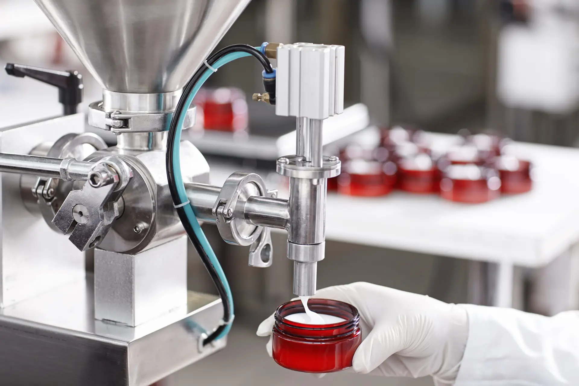 Hard mechanism of packing ready beauty cream into red container. A factory worker dressed in white lab gown and gloves holding red container under tap pouring ready cream. Production process of cream Hard mechanism of packing ready beauty cream into red container. A factory worker dressed in white lab gown and gloves holding red container under tap pouring ready cream. Production process of cream